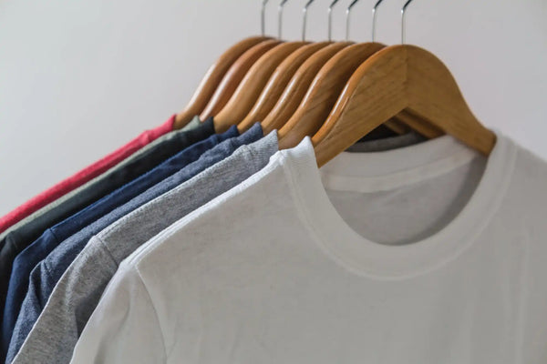 White crew neck t-shirt on wooden hanger with multiple colors of shirts behind it.